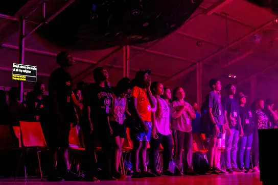Campers on their feet in a dark hall lit with purple stage lights